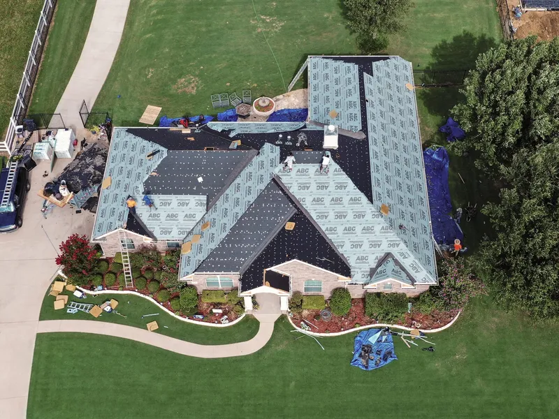 Overhead view of a large brick home mid-replacement, with most roof slopes stripped to the decking and covered in synthetic underlayment, shingle bundles staged on the ridges and tarps protecting the landscaping below.
